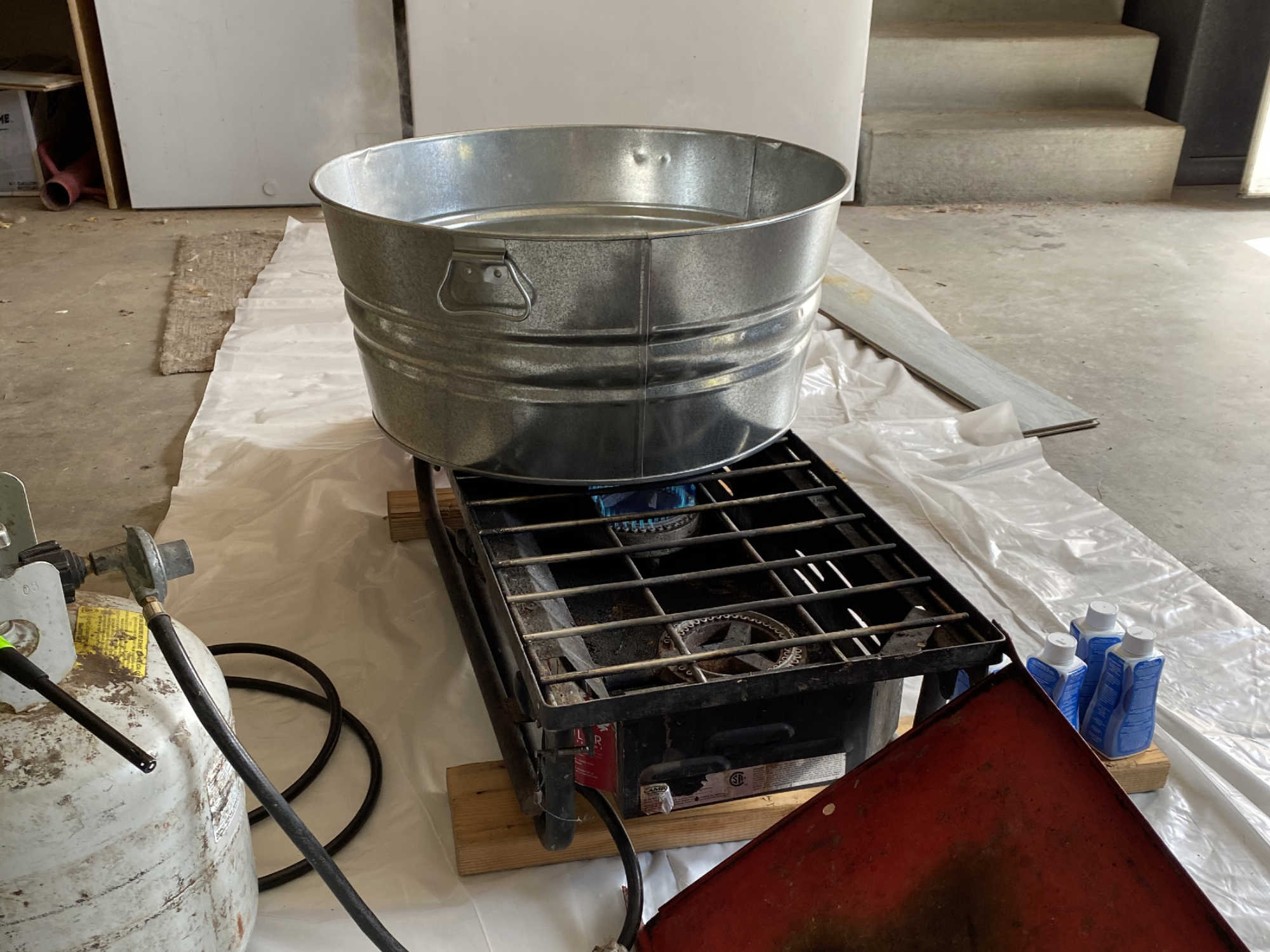 A metal tub full of boiling water, being heated by a propane camp stove in a garage. The floor is covered in a tarp and a cooking thermometer has been inserted in the water to ensure the right temperature.
